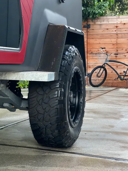 Rugged RV rental: close-up of its large off-road tire & suspension. Vehicle parked on wet pavement by a wooden fence & bike.. Escapod Escapod Trailer 2023