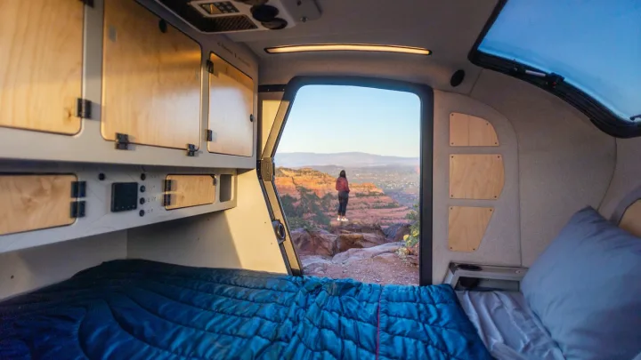 Wake up to adventure with an RV rental! Inside a cozy camper, a bed looks out to a woman on a cliff, red rock landscape.. Escapod Escapod Trailer 2023