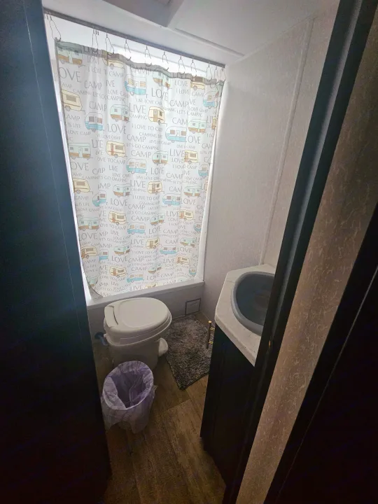 Compact RV rental bathroom features a shower with camping-themed curtain, toilet, and sink, designed for travel.. Forest River Cherokee Grey Wolf Special Edition 2022