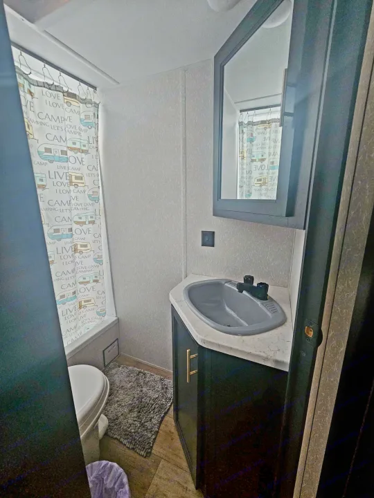 Inside an RV rental: a compact bathroom with a grey corner sink, white toilet, and a "Love Camping" themed shower curtain.. Forest River Cherokee Grey Wolf Special Edition 2022