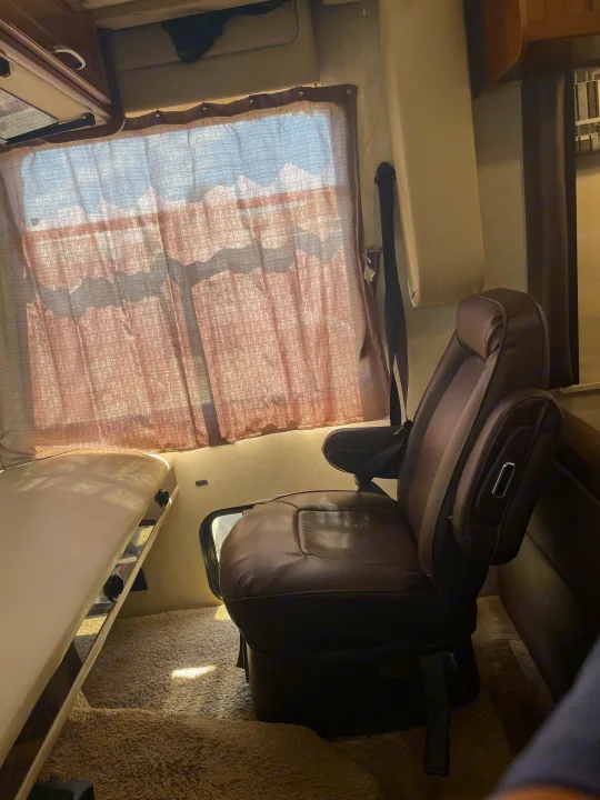 RV rental interior view: brown captain's chair, sunlit window with sheer curtains, and a beige narrow bed/bench.. Coachmen Pursuit 2014