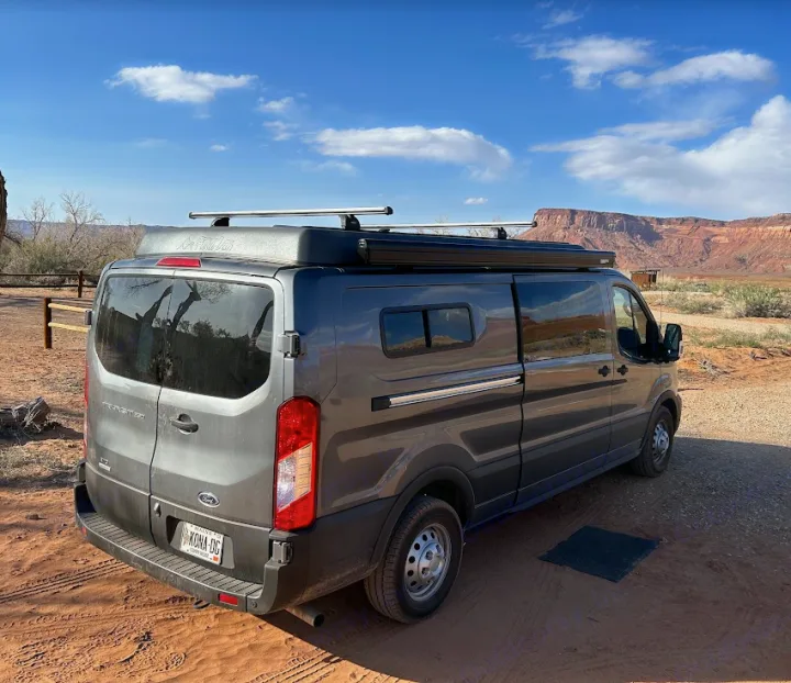 Gray Ford Transit campervan (rv rental) with rooftop tent & awning in a sunny red rock desert. Perfect for outdoor adventure.. Ford Custom Transit 2022