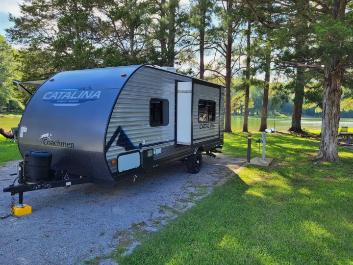 This RV rental, a travel trailer, is set up at a serene lakeside campsite among tall trees. Perfect outdoor escape.. Coachmen Catalina Summit Series 7 2023