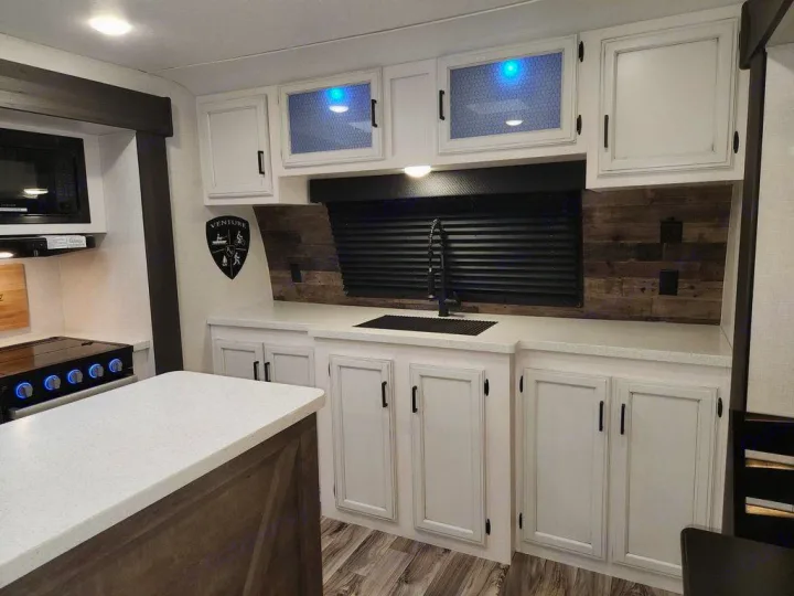 This modern RV rental kitchen features white cabinets, a sink, microwave, and stove, with a stylish wood backsplash.. SportTrek STT333VFK 2023
