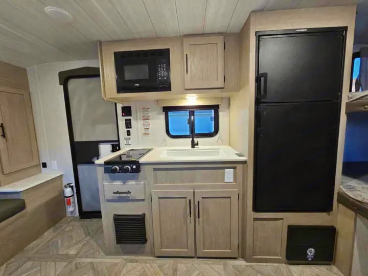 This compact rv rental kitchen features light wood-look cabinets, microwave, sink, two-burner stove, and fridge, optimizing space for travel.. Forest River Salem Cruise Lite 2024