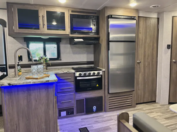 Bright RV rental kitchen featuring stainless steel appliances, a modern sink, wood cabinets, and cool blue accent lighting.. Crossroads RV Zinger Lite 2022