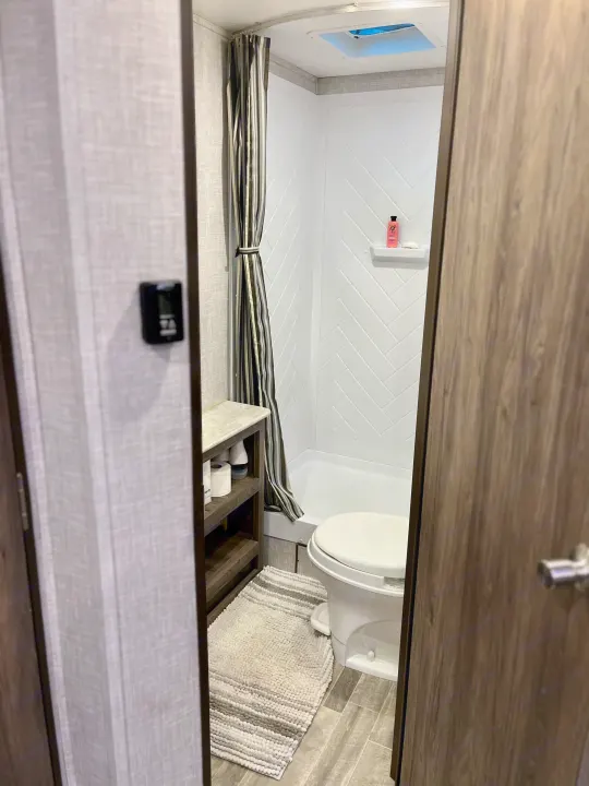 RV rental bathroom features a compact shower with striped curtain, toilet, and wood-look storage shelving.. Crossroads RV Zinger Lite 2022