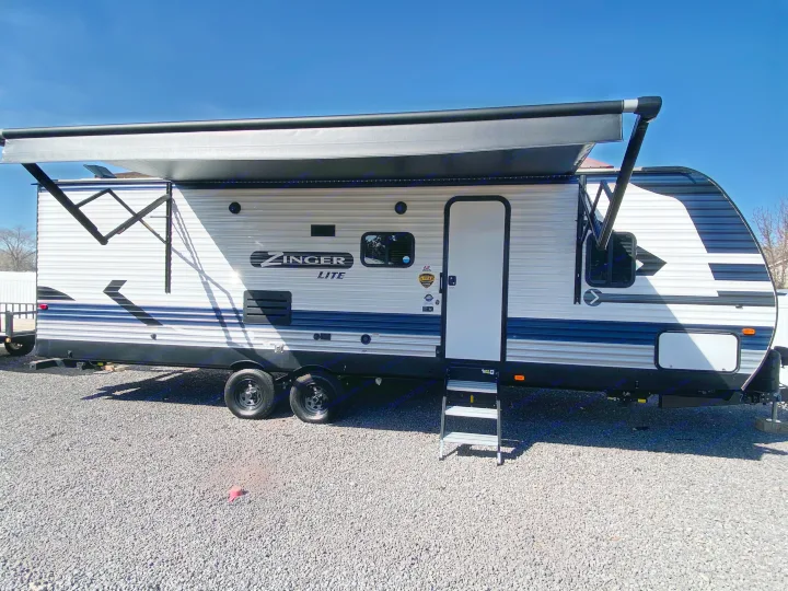 Exterior view of a travel trailer RV rental with extended awning & entry steps on a gravel lot under a clear sky.. Crossroads RV Zinger Lite 2022