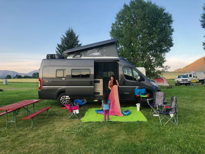 Family preparing camp with their **RV rental**, a gray camper van with pop-top, at a grassy campsite.. Winnebago Solis 2023