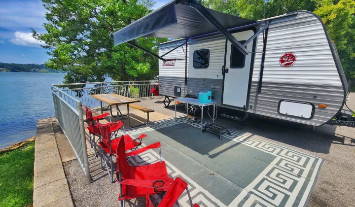 RV rental by the lake, featuring an extended awning, outdoor rug, multiple red chairs, and a picnic table for relaxation.. Coleman Lantern 2023