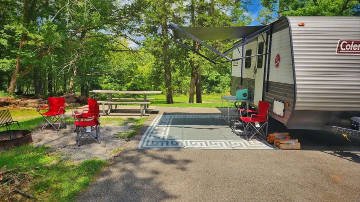 Scenic RV rental campsite. Features travel trailer with awning, red chairs, picnic table, and fire pit in a natural setting.. Coleman Lantern 2023