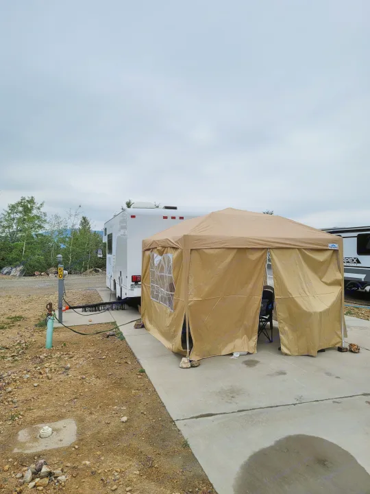 An RV rental parked at a campsite next to a tan canopy, with utility hookups and mountain views under a cloudy sky.. Thor Majestic 2017