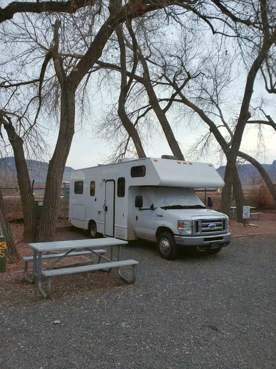 White RV rental parked at a winter campsite with a picnic table and bare trees. Mountains are visible in the background.. Thor Majestic 2017