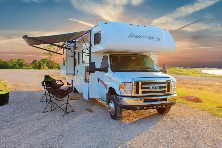 This white RV rental, a Forest River Sunseeker, is parked at a gravel campsite with its awning extended and chairs ready for outdoor relaxation.. Forest River Sunseeker 2015