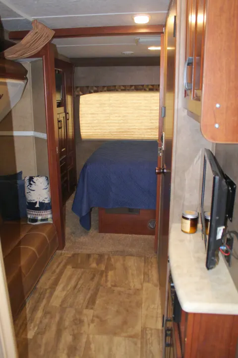 This RV rental features a compact living area with a sofa, leading to a cozy bedroom with a blue bedspread and window. Ideal for travel.. Forest River Sunseeker 2015