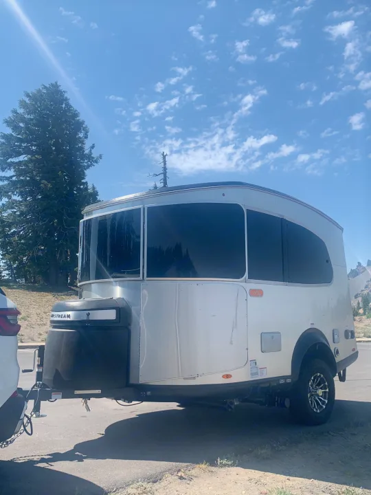 This Airstream Basecamp RV trailer, ideal for an rv rental adventure, shines under a sunny sky with trees and mountains.. Airstream Basecamp 2022