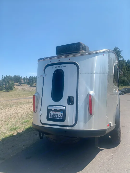 A silver Airstream travel trailer (CA plate 1NW9122), an RV rental, parked on asphalt with forested hills and clear skies behind it.. Airstream Basecamp 2022