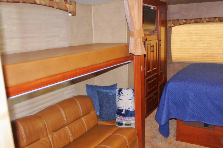 Spacious RV rental interior: lower bunk with brown couch, upper bunk, main bed with blue quilt, and wooden storage cabinets.. Forest River Sunseeker 2015