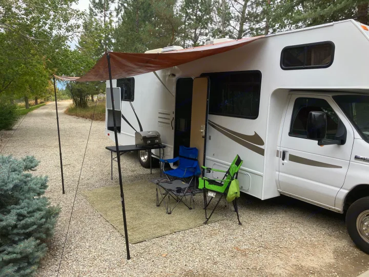 An RV rental parked on gravel with an outdoor living setup: awning, grill, and chairs for a relaxing campsite.. Majestic Ford 2018