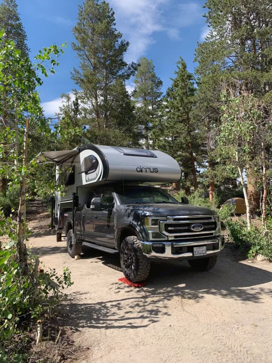 A grey Ford truck equipped with a silver Cirrus truck camper is parked on a dirt road in a sunny, tree-lined campsite, perfect for an RV rental getaway.. Ford F350 Diesel Double Cab 2020