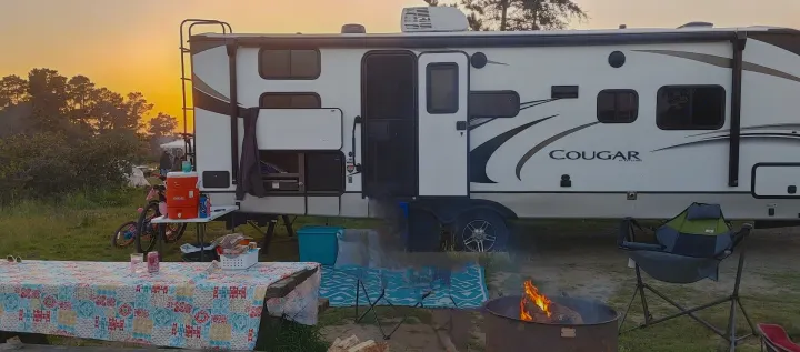 Enjoying an RV rental adventure: a travel trailer parked in a grassy camp, with a campfire burning, picnic table, and open outdoor kitchen.. Keystone RV Cougar 2021