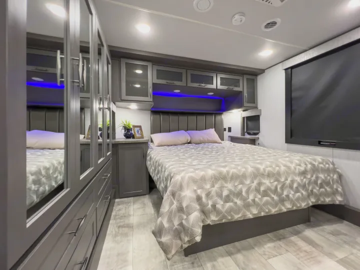 Inside this modern RV rental bedroom: a large bed, overhead cabinets with blue accent lights, and mirrored closet doors.. Grand Design Momentum 2023