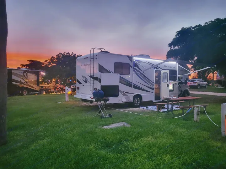 A bright RV rental at sunset in a picturesque campground. Its awning and interior lights create a welcoming ambiance.. Thor Four Winds 2021