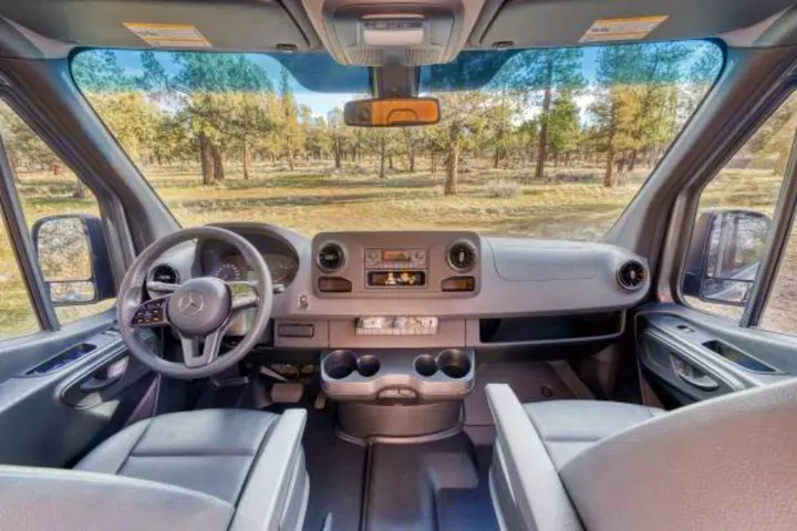 View from an RV rental's spacious driver seat, showing the steering wheel, dashboard, and a natural forest landscape outside.. Mercedes-Benz Sprinter 2021