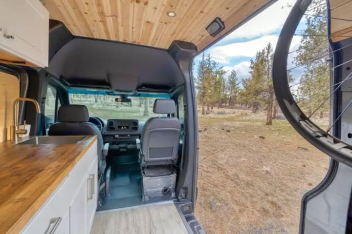 RV rental camper van interior with kitchen, front seats, and a bike, overlooking an outdoor forest landscape.. Mercedes-Benz Sprinter 2021