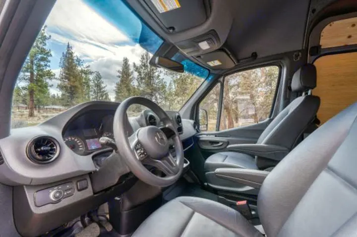 Inside an RV rental van: the spacious front cabin with dashboard, steering wheel, and seats, overlooking a forest.. Mercedes-Benz Sprinter 2021