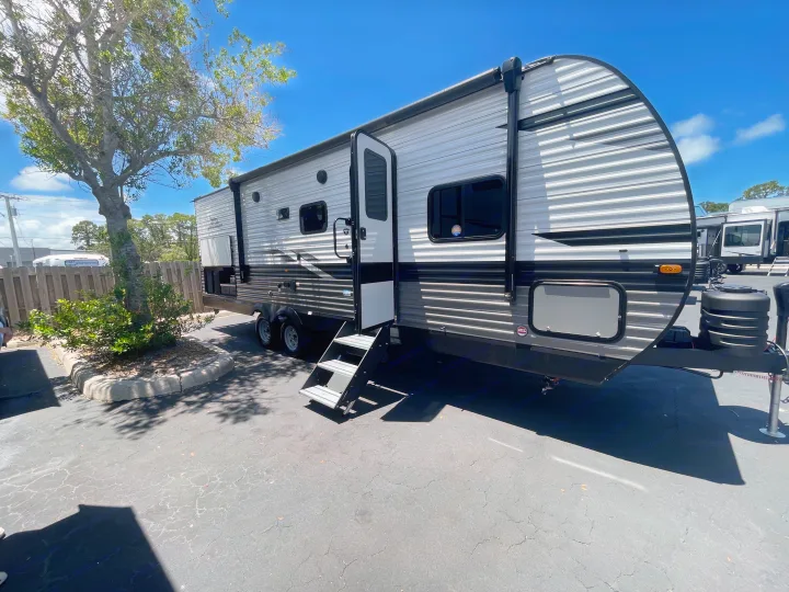 A clean, modern RV rental travel trailer with an open door and steps is parked on asphalt under a blue sky.. Jayco Jay Flight 2023