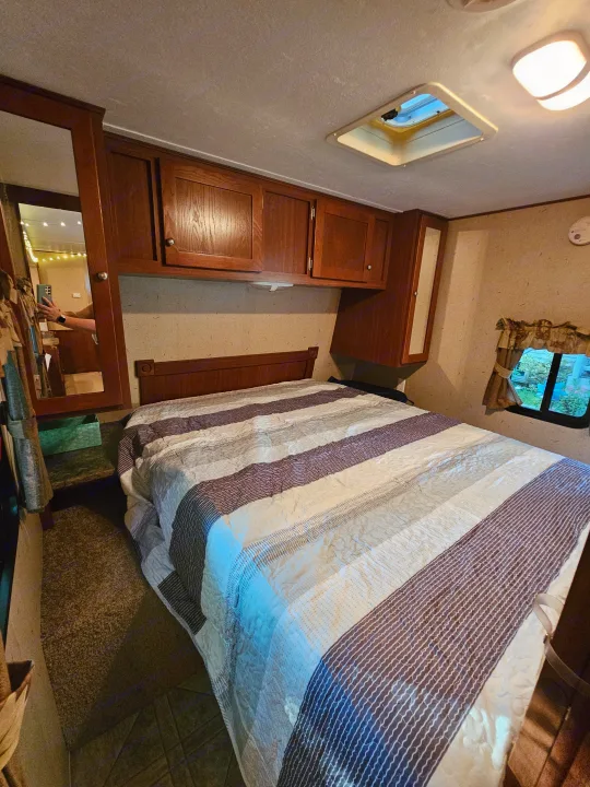 Cozy RV rental bedroom featuring striped bedding, overhead storage cabinets, a mirror, and a window with outdoor view.. Crossroads RV Zinger 2010