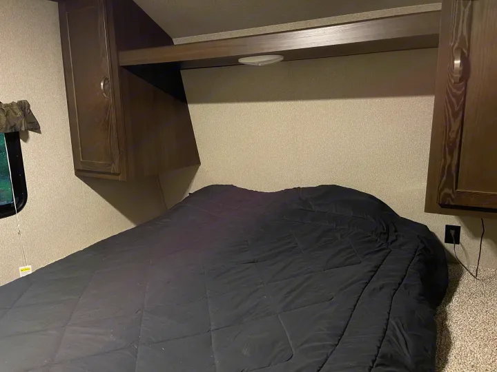 Bedroom interior of an RV rental, showing a bed with a dark comforter, overhead cabinets, and a window.. Keystone RV Springdale 2018
