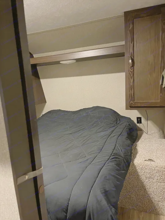 This RV rental's cozy bedroom features a bed with a black comforter, an overhead cabinet, and a convenient power outlet, optimized for compact sleeping.. Keystone RV Springdale 2018
