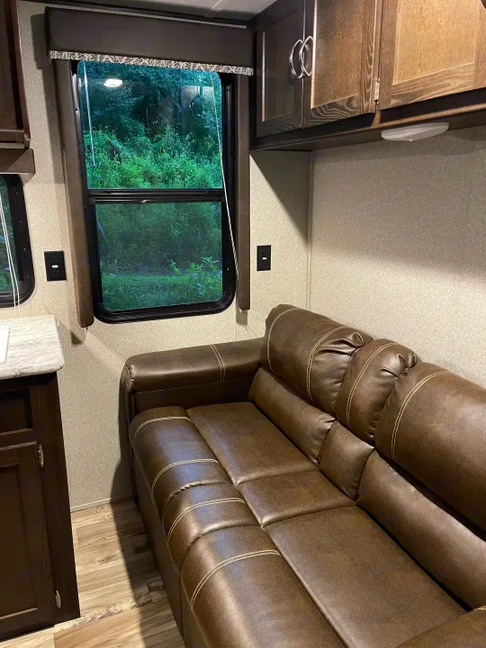 RV rental's inviting living space: brown sofa, window framing lush trees, and overhead cabinets. Cozy camper interior.. Keystone RV Springdale 2018