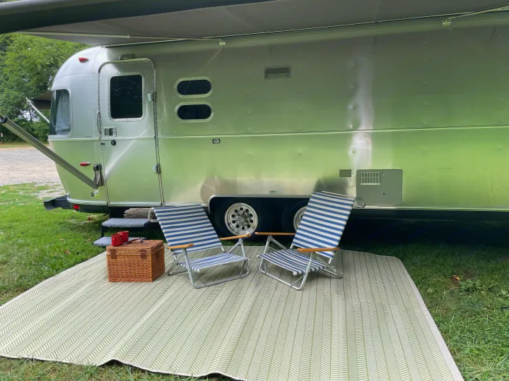 Outdoor living with an Airstream RV rental. An awning extends over striped chairs, picnic basket, and patterned mat on grass.. Airstream International 2014