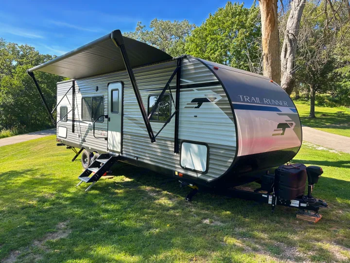 This modern travel trailer RV rental is set up in a sunny, treed park with its awning extended for shade.. Heartland RVs Trail Runner 2022