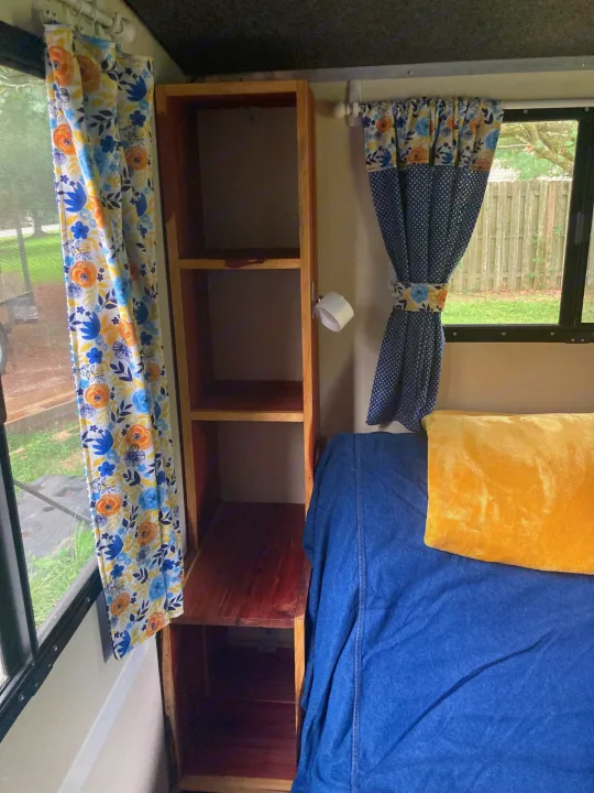 Inside an RV rental: floral window curtains, wooden shelves, and a cozy bed with blue and yellow bedding.. Runaway Rouser 2023