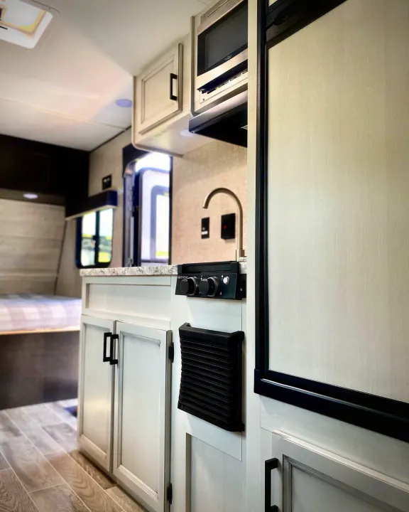 An RV rental's well-equipped kitchen featuring a stovetop, microwave, and refrigerator. Bedroom visible behind.. Jayco Jay Flight SLX 2022