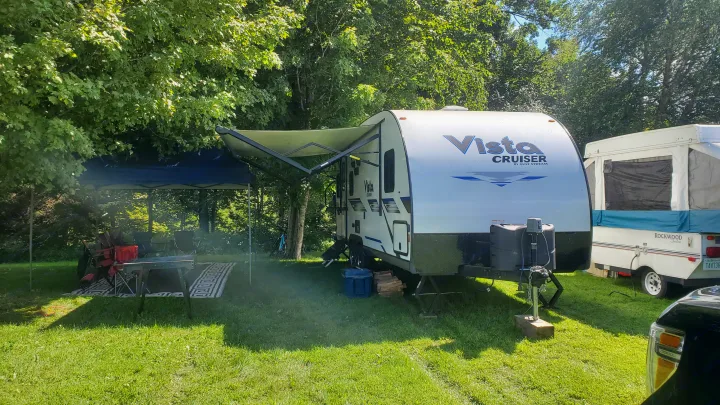 An RV rental campsite with a white Vista Cruiser travel trailer, extended awning, and a pop-up camper under shady trees.. Gulf Stream Vista Cruiser 2021