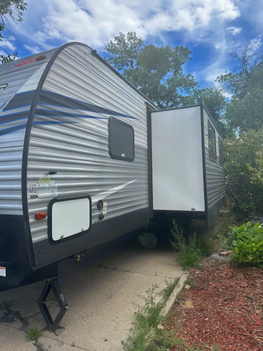 An RV rental, a grey travel trailer with a large slide-out, is parked on a paved surface, surrounded by greenery.. Avenger Avenger Trailer 2021