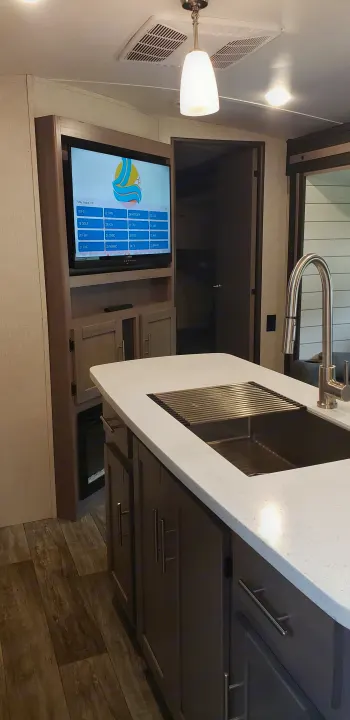 RV rental interior: kitchen island with sink, TV showing channel guide. A modern, functional space for travel.. Keystone Sunset Trail 2022