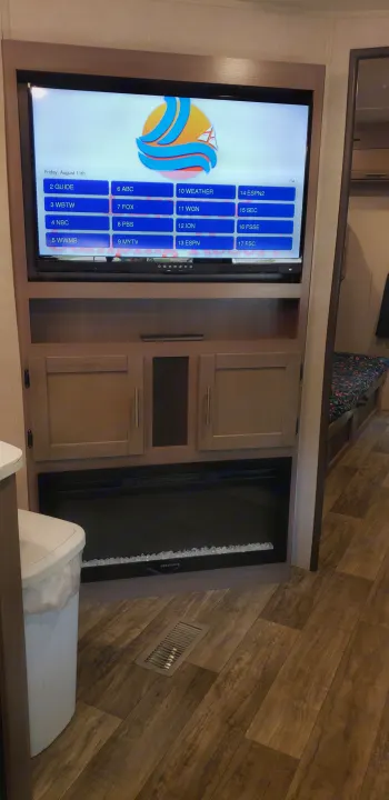 RV rental interior: TV displaying channel guide for Fri, Aug 11th, with cabinets & a Greystone fireplace below.. Keystone Sunset Trail 2022
