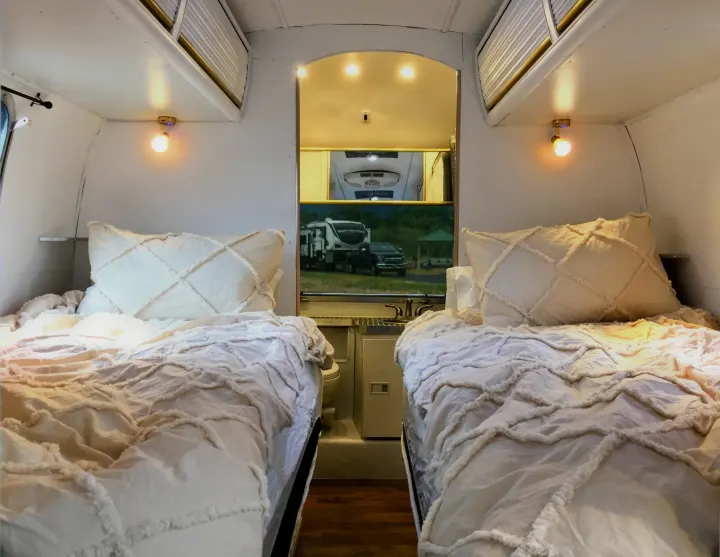 Relaxing RV rental interior with two twin beds featuring textured cream bedding. Includes a view of the bathroom/kitchenette area.. Airstream Trade Wind 1976