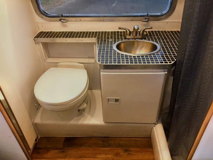Inside an RV rental: a compact bathroom featuring a white toilet, small tiled sink, and dark shower curtain.. Airstream Trade Wind 1976