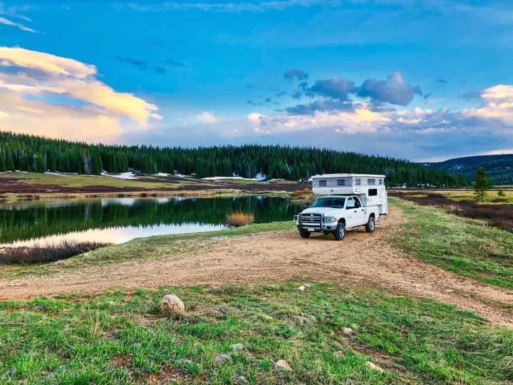 Experience nature with an RV rental! White truck camper by a calm lake, lush forest, and mountains under a vibrant sky.. Hallmark Everest 2011