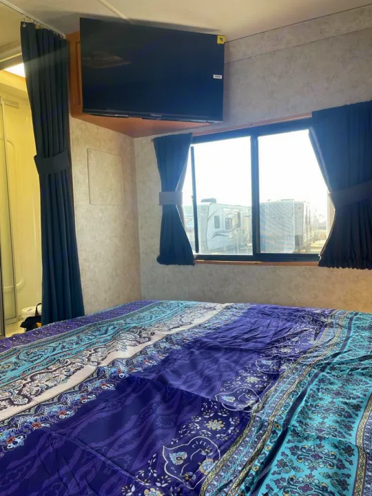 Comfortable RV rental bedroom featuring a purple and teal patterned comforter, wall-mounted TV, and window view.. Four Winds Majestic 2006