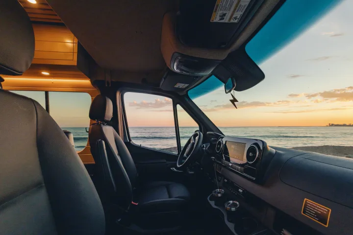 Experience coastal travel in an rv rental! Interior view of dashboard & seats, bathed in warm light, overlooking an ocean sunset.. Mercedes-Benz Sprinter 2023