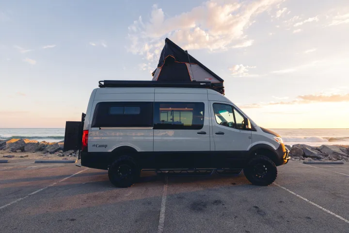 This modern camper van with a rooftop tent is parked by the ocean at sunset. An ideal rv rental for your next beach adventure.. Mercedes-Benz Sprinter 2023