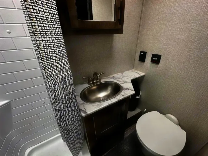 RV rental's compact bathroom features a shower with patterned curtain, sink, wood vanity, toilet, and wall controls.. Venture Rv Sonic 2022
